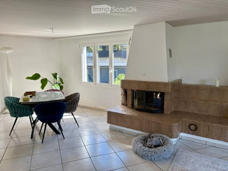 Bright living room with a fireplace, dining table, chairs, and a dog bed. The room features large windows and a modern design.