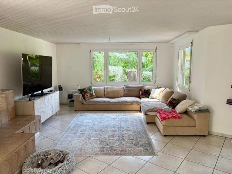Bright living room with large windows offering a view, featuring a sectional sofa, coffee table, television, and a decorative rug.