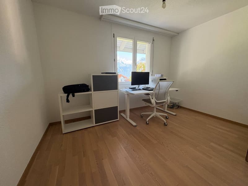 A modern, well-lit room with wooden flooring, a white desk, a chair, a screen, a keyboard, and a shelf with a bag on it.