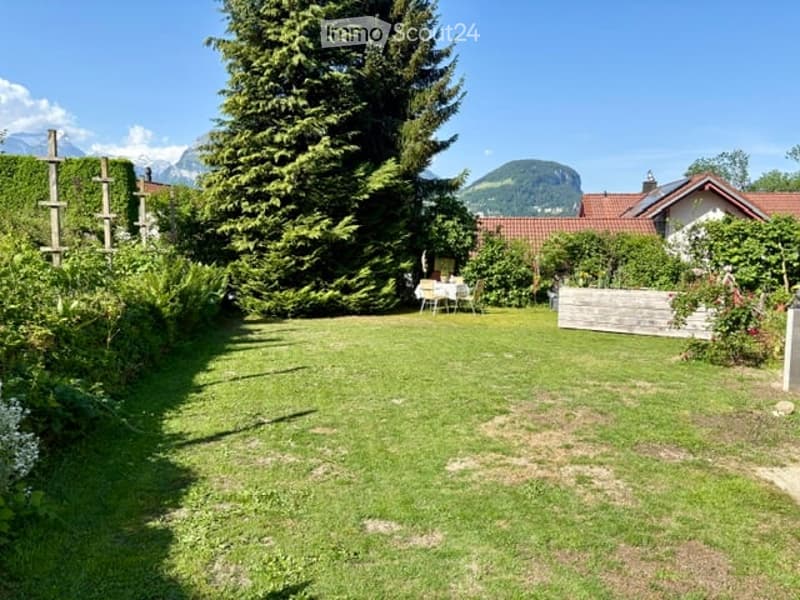 A well-maintained garden with a lawn, tall trees, a small patio area with a table and chairs, and a wooden fence. The background features a house with a red-tiled roof and distant mountain views.