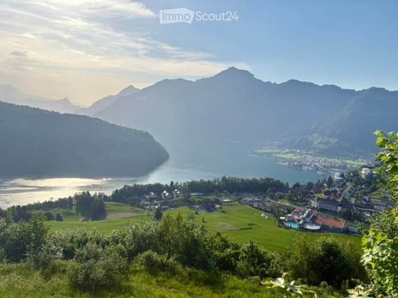 Panoramic view of a lake surrounded by mountains, with a town and green fields in the foreground.