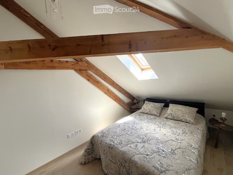 Cozy attic bedroom with wooden beams, skylight window, bed with patterned bedding, nightstand, and table lamp.