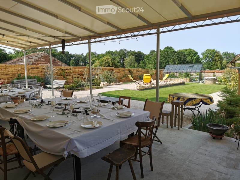 An outdoor dining area with a long table set for a meal, surrounded by chairs, a smaller table, and a garden with lawn, plants, and lounge chairs.