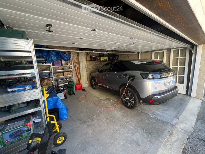 Garage with a parked car, shelves with tools and storage items, and a ladder. The garage door is open, revealing the interior space.