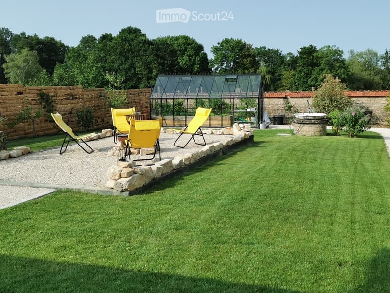 Well-maintained garden with a lawn, stone border, yellow lounge chairs, a greenhouse, and a stone fountain.