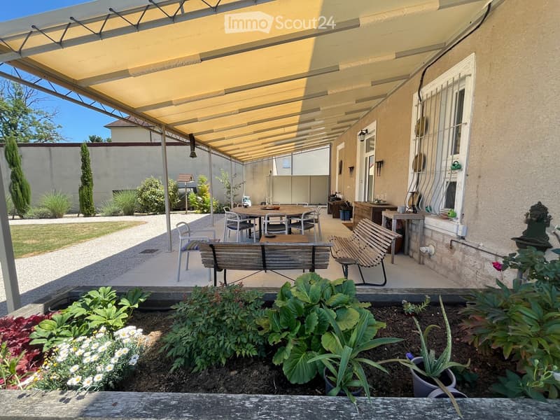A covered outdoor patio area with a large table, chairs, benches, and surrounding greenery, including potted plants and flowers.