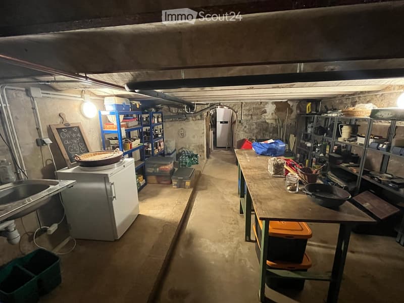 A basement or utility room with concrete walls, shelves, a sink, a refrigerator, a table with a frying pan, and various storage items.