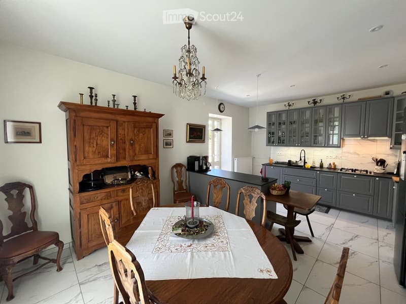 A well-furnished room with a wooden dining table, chairs, a large wooden cabinet, modern kitchen cabinets, and a chandelier.