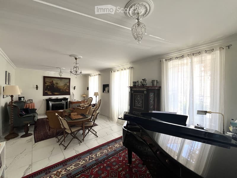 Elegant living room with a grand piano, fireplace, chandelier, and large windows with curtains. Features a mix of seating including a sofa, armchairs, and a dining table with chairs.