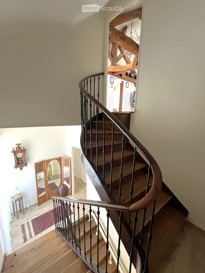 Spiral staircase with wooden steps and metal railing, light-colored walls, wooden floor, small table, mirror, and clock visible in the background.