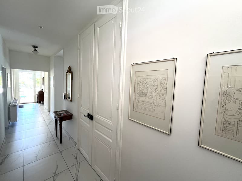 A bright, white-walled hallway with a small table, mirror, and two framed artworks. The hallway leads to an open area with a swimming pool visible in the background.