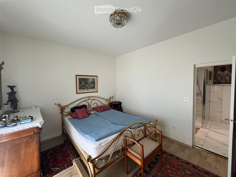 A furnished bedroom with a metal-framed bed, blue and red bedding, a wooden nightstand, a small wooden chair, a chandelier, and a picture frame on the wall. A small table with various items is visible on the left side.