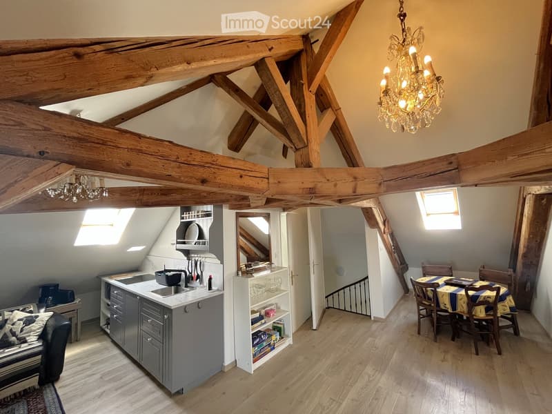 Loft-style room with exposed wooden beams, a modern kitchen area, a dining table with chairs, and a cozy seating area with a sofa.