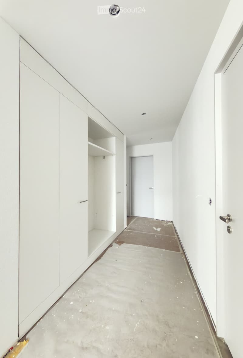 Empty hallway with white walls, concrete floor, and built-in white cabinetry on one side. A closed door is visible at the end of the hallway.