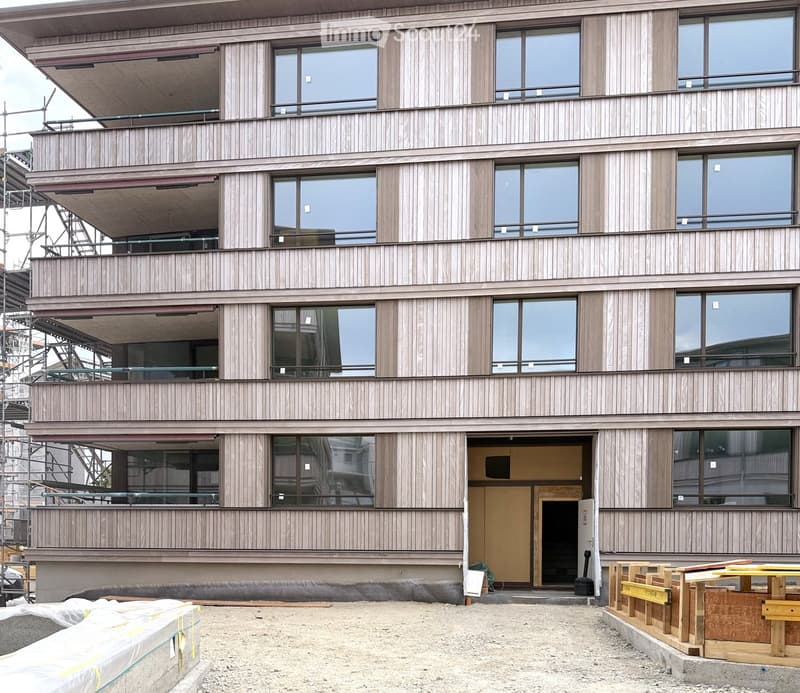 Modern multi-story building with wooden siding and large windows, featuring balconies and a partially constructed entrance area with scaffolding and construction materials.