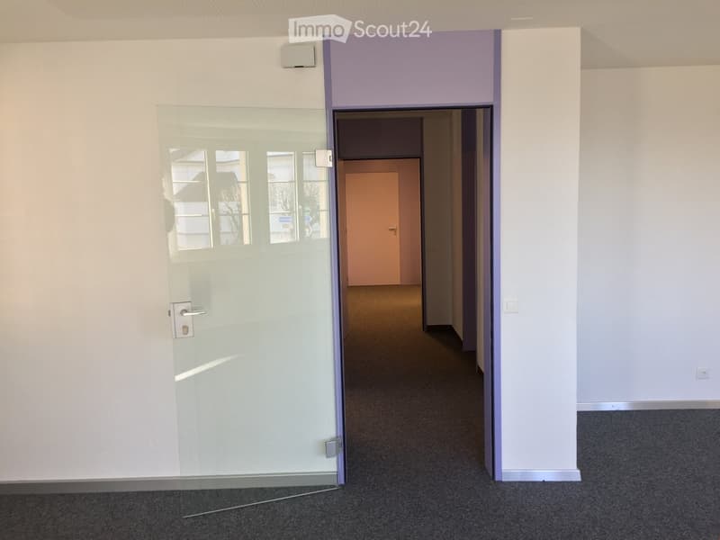 A room with a glass door leading to a hallway, the walls are white and the floor is carpeted, with a visible door at the end of the hallway.