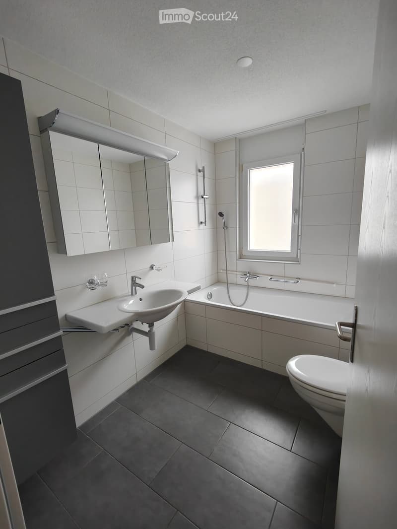 Modern bathroom with white tiles, dark grey floor, sink, bathtub, toilet, mirror, and showerhead.