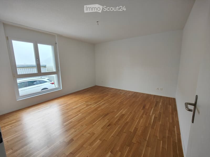 Empty room with wooden flooring, white walls, and large windows with a view of a parked car outside.