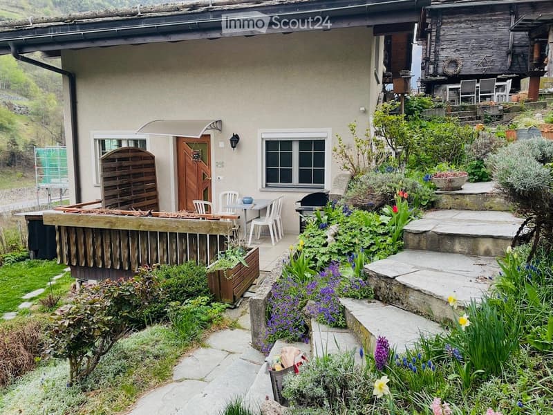 A well-maintained garden with a stone pathway, colorful flowers, shrubs, and a small seating area with white plastic chairs and a table. The house features a wooden door, a window, and a wooden fence.
