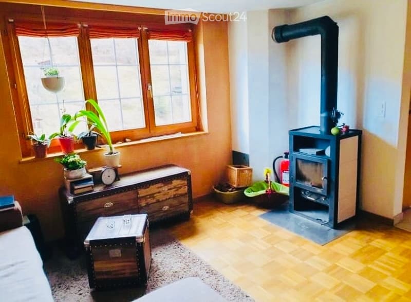 Cozy living room with wooden flooring, large windows, fireplace, sofa, wooden cabinet, and potted plants on the windowsill.