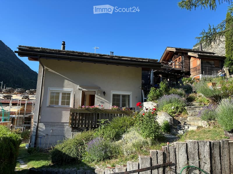A two-story house with a wooden balcony, surrounded by a garden with flowers and shrubs, set against a mountainous backdrop.