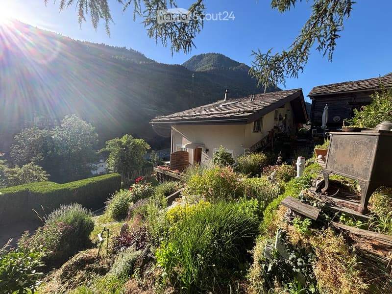 A picturesque garden area with a house in the background, surrounded by lush greenery, flowers, and a wooden fence. The house has a stone facade and a sloped roof, with a staircase leading up to the entrance.