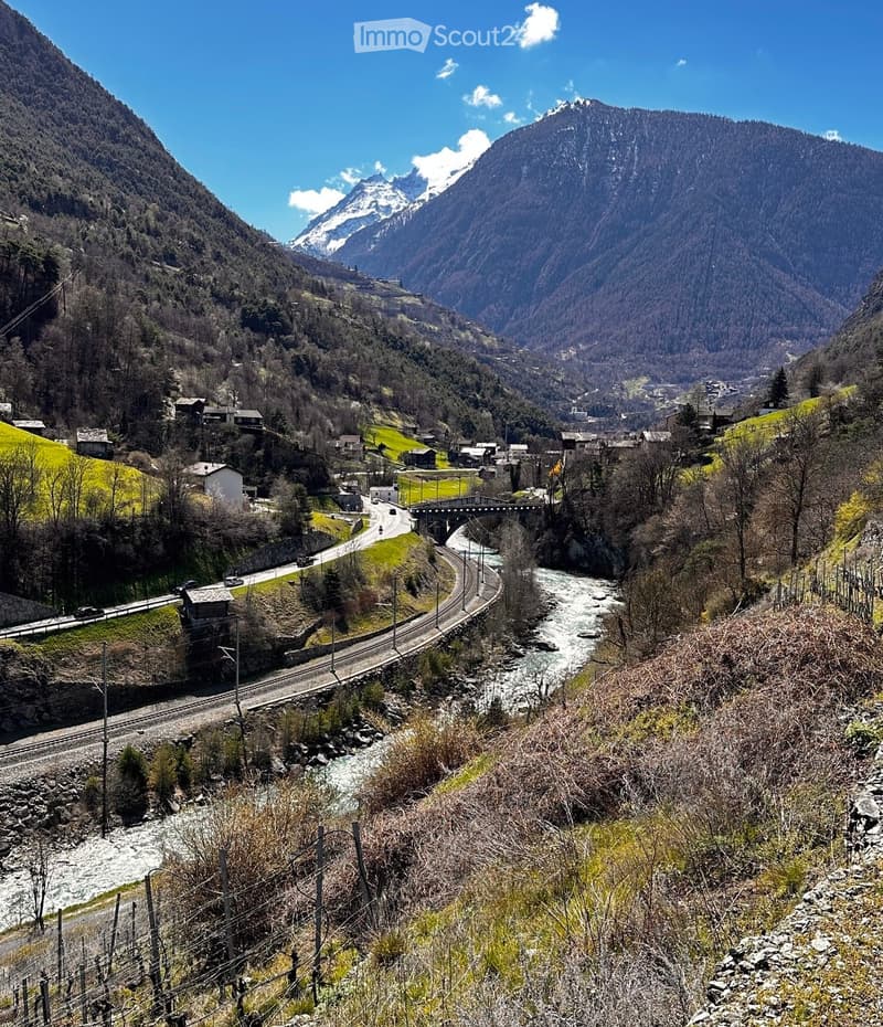 A scenic mountain valley with a river, small village, and winding road surrounded by snow-capped peaks and green fields.
