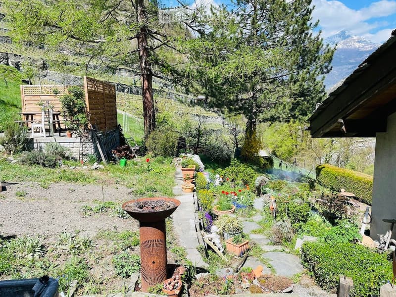 A well-maintained garden with a stone pathway, various potted plants, a metal fire bowl, and a wooden deck area with benches and a table. The garden is surrounded by trees and offers a scenic mountain view in the background.