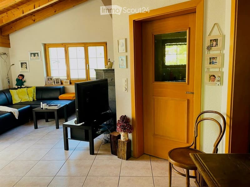 Living room with a black leather sofa, coffee table, television, wooden chair, and a small table with a vase of flowers. The room features large windows, picture frames on the walls, and a cozy, inviting atmosphere.