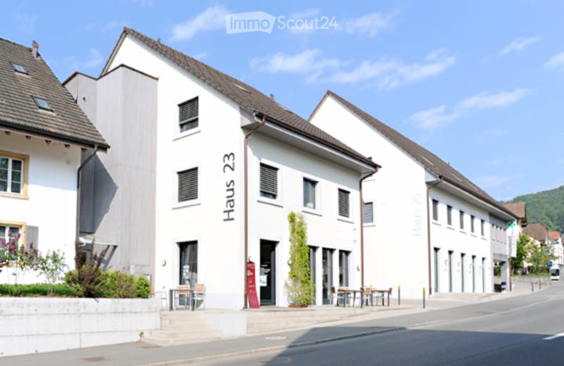 White exterior, brown roof, multiple windows, outdoor seating, tree, number 23 on the front