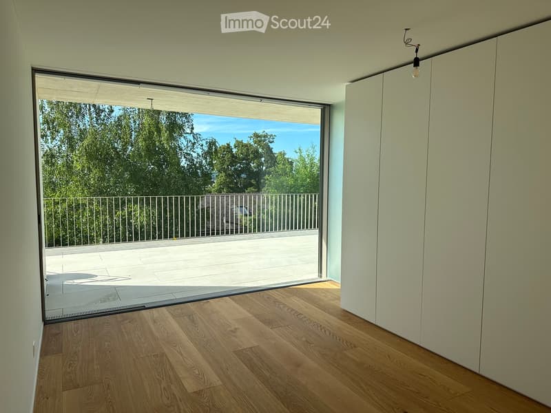 Empty room with wooden flooring, large sliding glass door leading to a balcony with a metal railing, and a view of greenery outside.