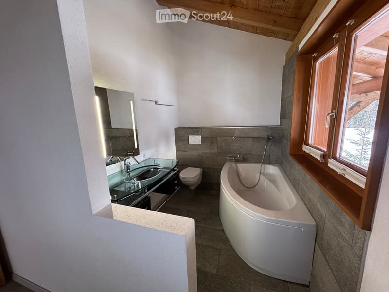 Modern bathroom with a bathtub, toilet, sink, and mirror. Features wooden beams on the ceiling and large windows with wooden frames.