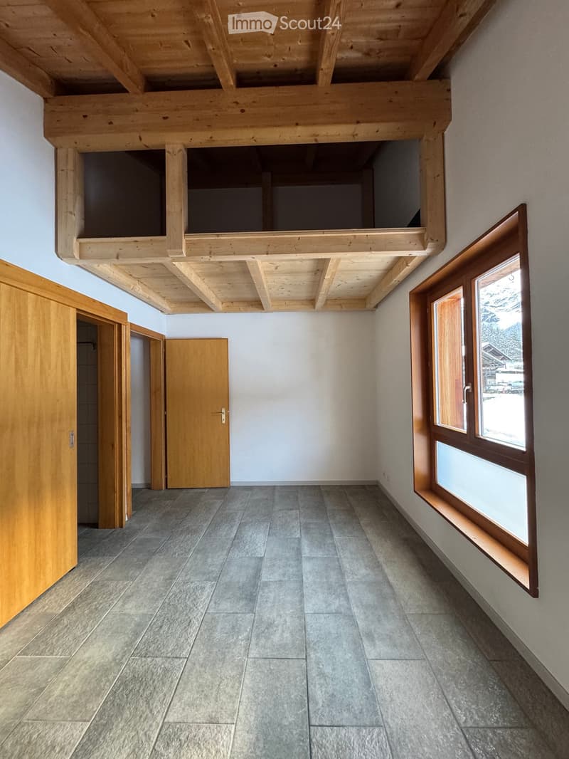 Empty room with wooden ceiling beams, grey tiled floor, and large windows with wooden frames. The room has a wooden door and an open loft area above.