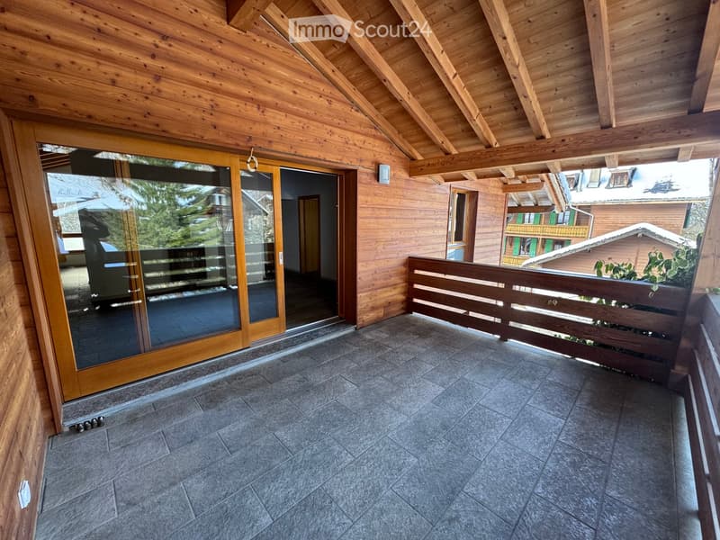 Wooden balcony with grey tiled floor, wooden railing, and large glass sliding doors leading to the interior.