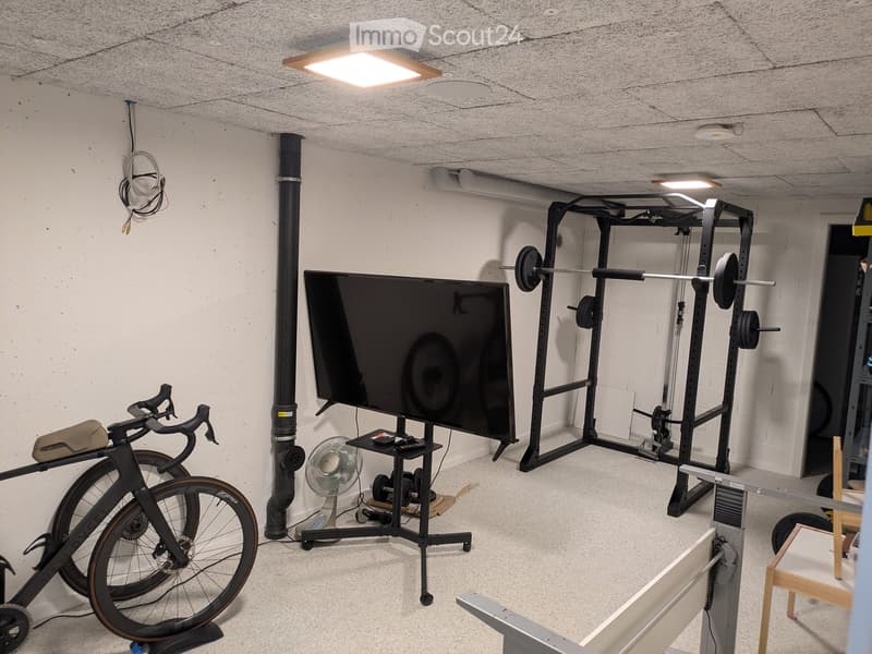 A well-lit room with a bicycle, TV, fan, weight machine, and a small stool.