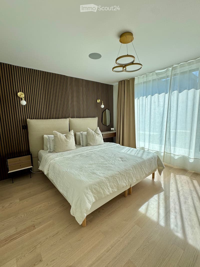 Modern bedroom with a large bed, nightstand, mirror, wall-mounted lamp, and pendant light. The room features wooden flooring and large windows with curtains.