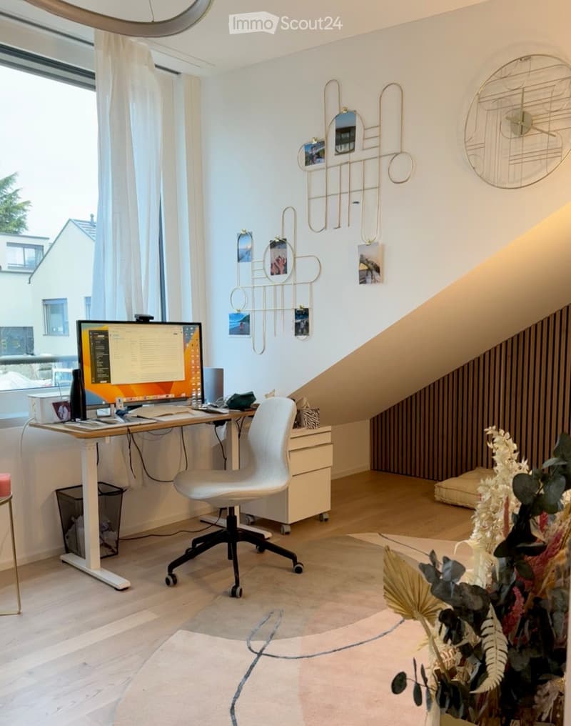 A modern, well-lit room with a desk setup, a white ergonomic chair, a large screen, and decorative wall art. The room features wooden flooring, large windows with curtains, and a cozy rug.