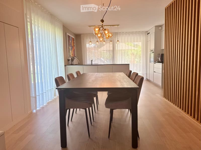 Modern dining area with a dark wooden dining table and six matching chairs, integrated kitchen with white cabinets and stainless steel appliances, large windows with sheer curtains, and a stylish pendant light fixture.