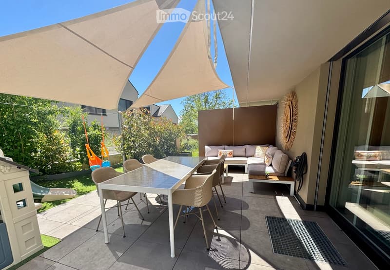 A modern outdoor patio with a large sunshade, a white dining table with four chairs, a sofa with cushions, and a small bench. The area is surrounded by greenery and has a view of the garden.