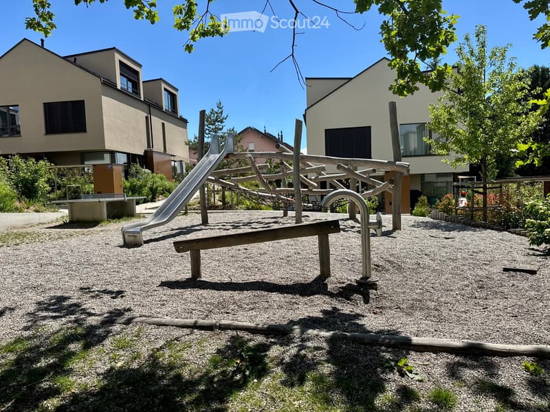 Playground area with a slide, seesaw, and climbing structure, surrounded by modern residential buildings and trees.