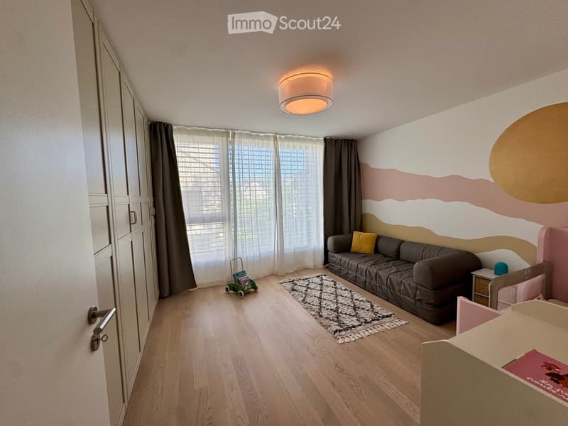 A cozy living room with a grey sofa, a patterned rug, a small toy car, a nightstand, and a bed with a colorful headboard.