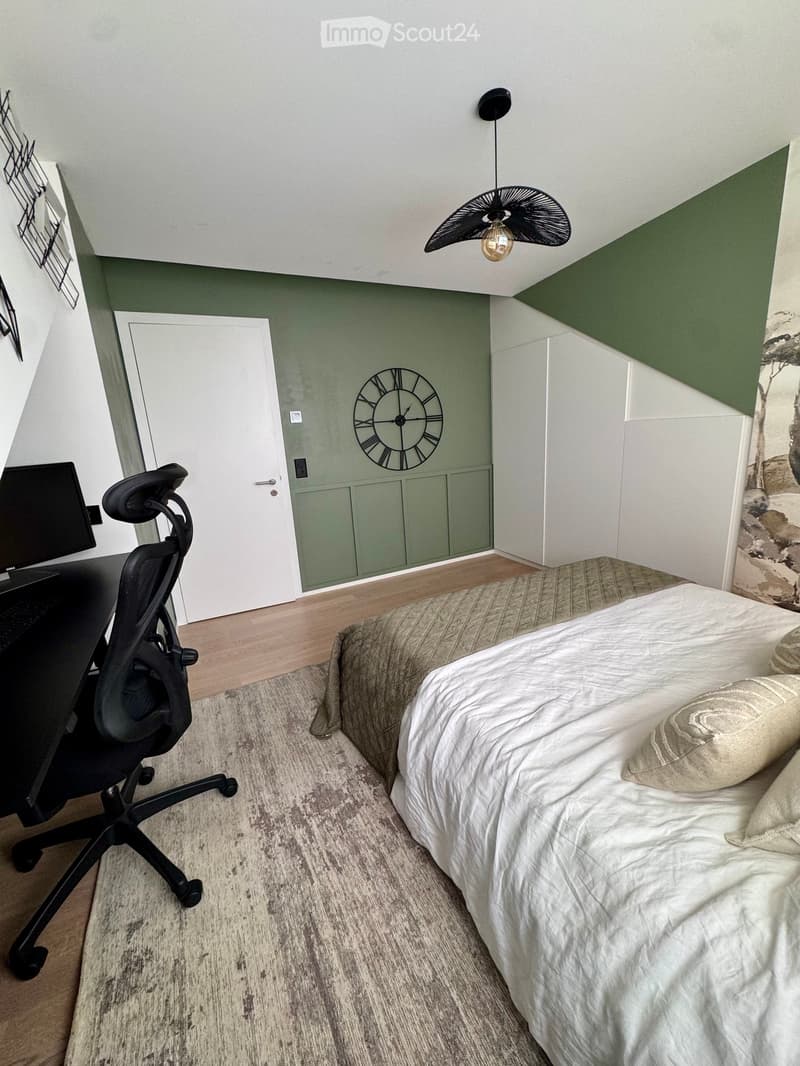 Cozy bedroom with a bed, desk, chair, and a stylish clock on the wall. The room features a slanted ceiling and modern decor.