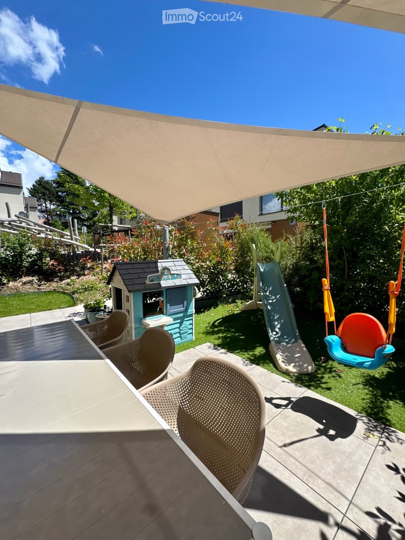 Outdoor patio with a table and chairs, a small playhouse, a slide, and a swing set, surrounded by greenery and a large shade sail.
