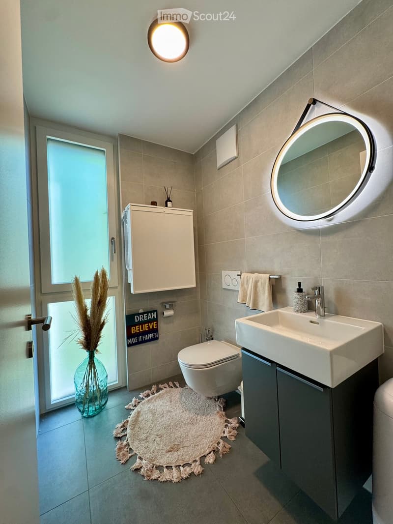 Modern bathroom with a white sink, toilet, round illuminated mirror, cabinet, towel rack, and a decorative vase with dried plants on the floor.
