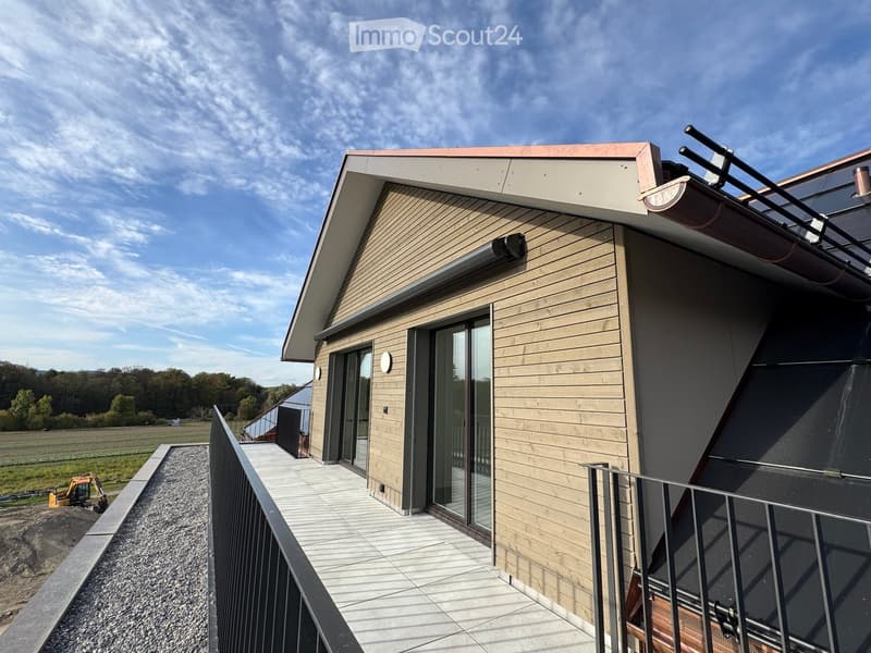 Modern house exterior with wooden siding, large glass windows, and a raised ground floor balcony overlooking a rural landscape.