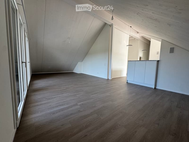 An empty, modern room with wooden flooring, white walls, and large windows. The space features a minimalist design with exposed wooden beams on the ceiling and a small kitchen area visible in the background.