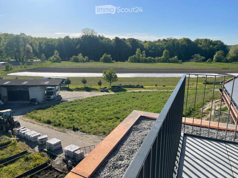 View from a balcony overlooking a large, open green area with scattered trees, a parked van, and construction materials. The foreground shows a metal railing and a paved area.