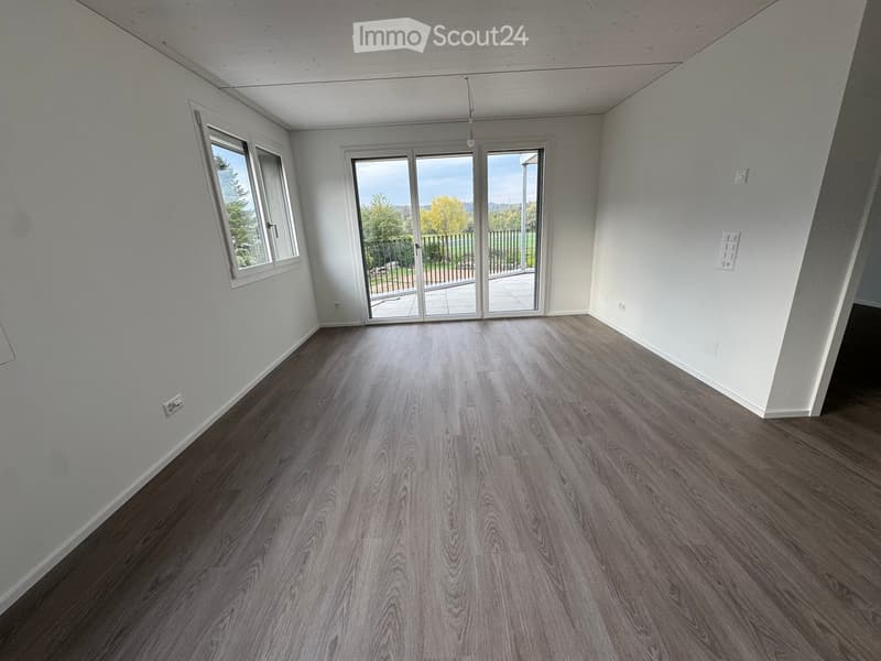 Empty room with wooden flooring, white walls, large windows, and sliding glass doors leading to a balcony.