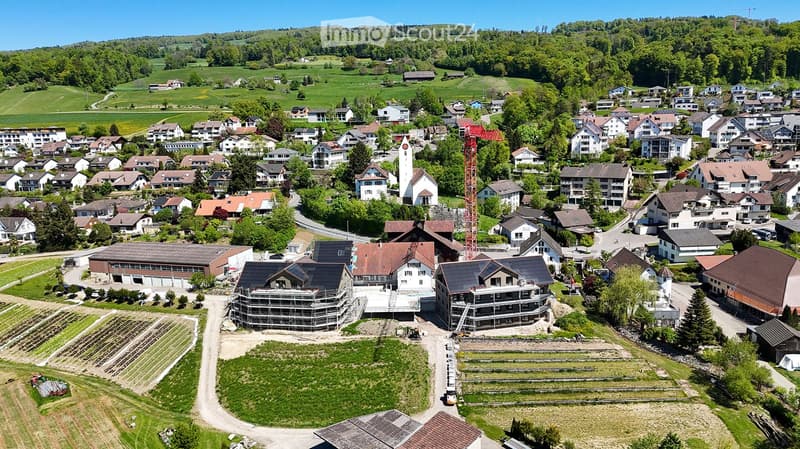 Aerial view of a small village with numerous houses, a church tower, and a construction site with scaffolding and cranes.