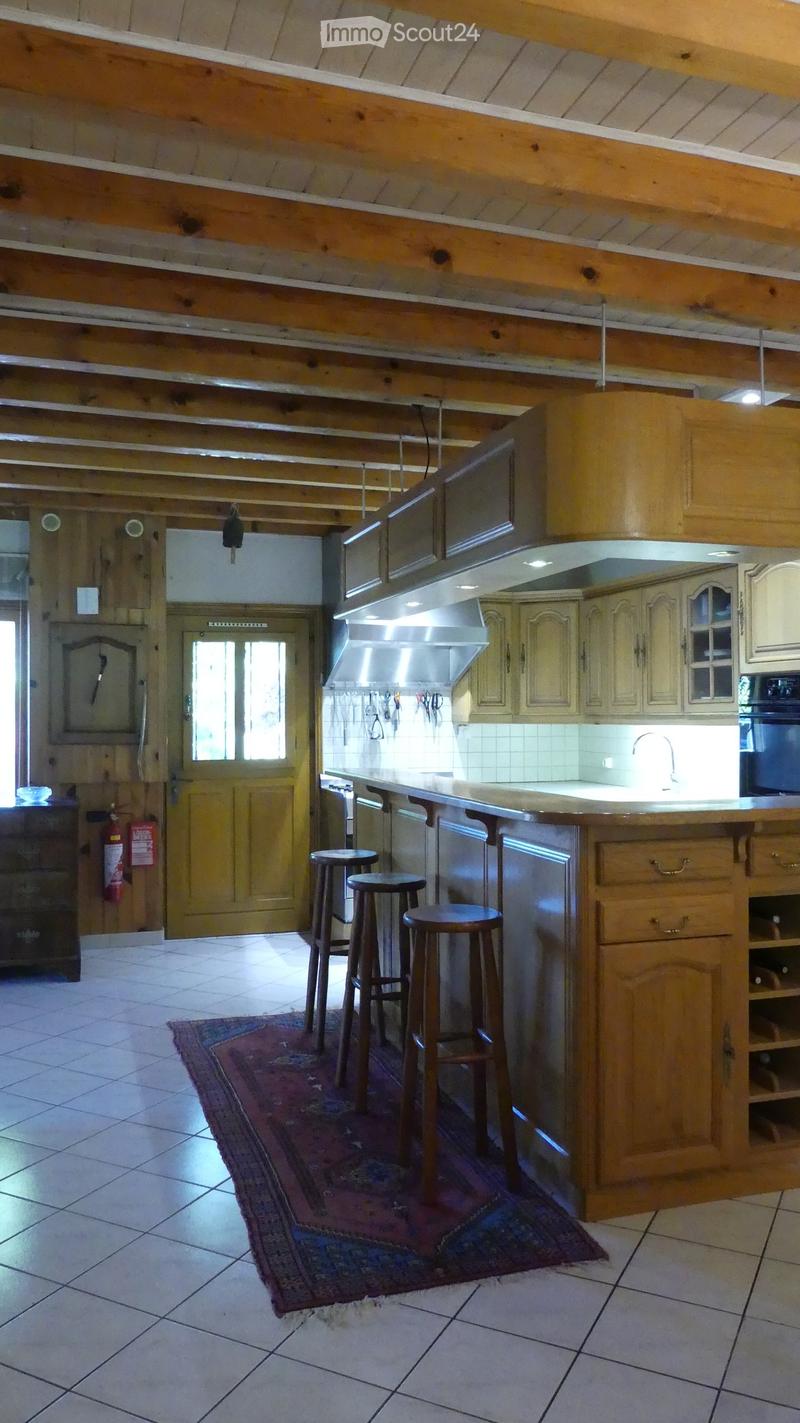 Bright kitchen with wooden beams, white tiled floor, and a central island with three wooden stools. The kitchen features light-colored cabinets, a stainless steel range hood, and a sink with a modern faucet.